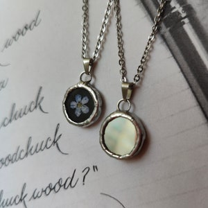 May include: Two silver necklaces with round pendants. One pendant features a pressed forget-me-not flower on a black background. The other pendant is white and iridescent.