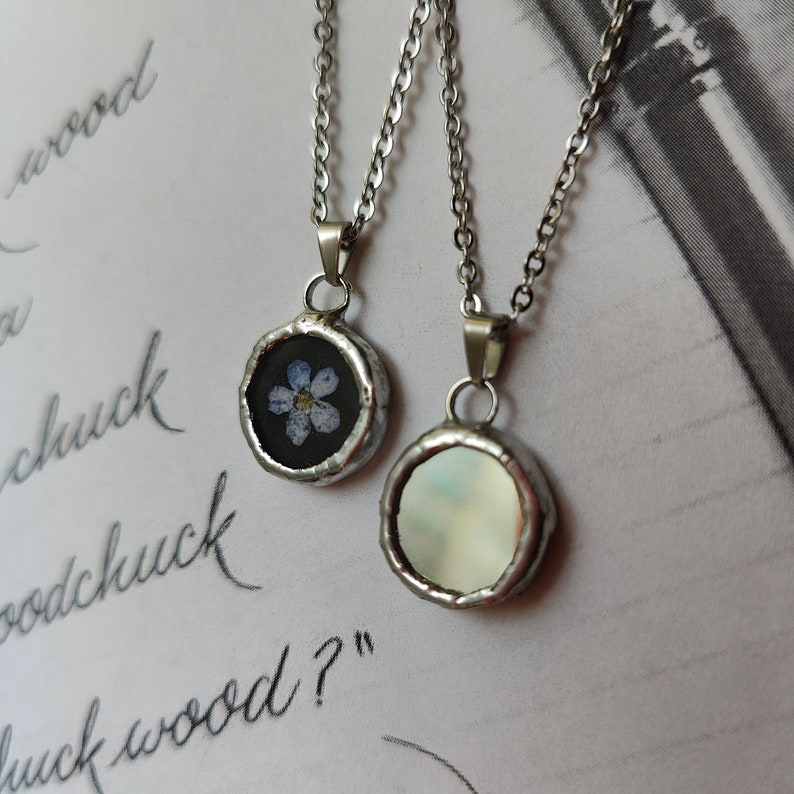 May include: Two silver necklaces with round pendants. One pendant features a pressed forget-me-not flower on a black background. The other pendant is white and iridescent.