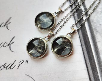 May include: Three silver-toned necklaces featuring round pendants. Each pendant contains a dark, circular background with white, wispy dandelion seeds encased under a clear, smooth, domed surface. The pendants hang from thin, matching silver-toned metal chains.