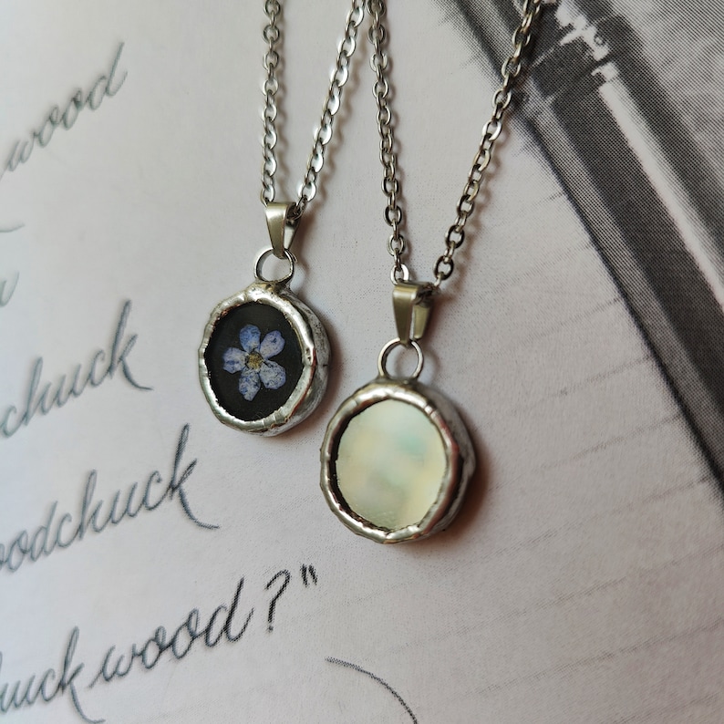 May include: Two silver necklaces with round pendants. One pendant features a pressed forget-me-not flower in a clear resin setting. The other pendant is a white iridescent shell.