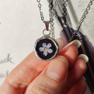 May include: A silver chain necklace with a round pendant featuring a pressed forget-me-not flower encased in resin.