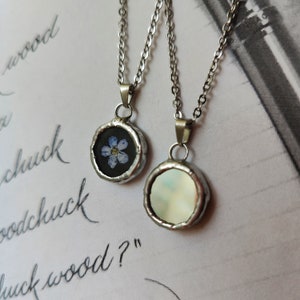 May include: Two silver necklaces with round pendants. One pendant features a pressed blue flower, the other has a white iridescent background.
