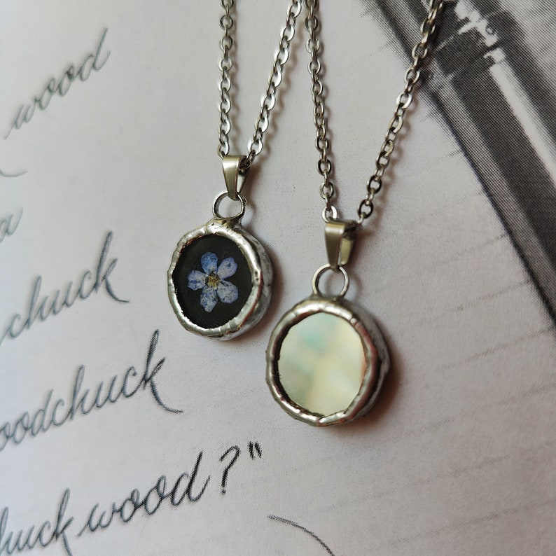 May include: Two silver necklaces with round pendants. One pendant features a pressed blue flower, the other has a white iridescent background.