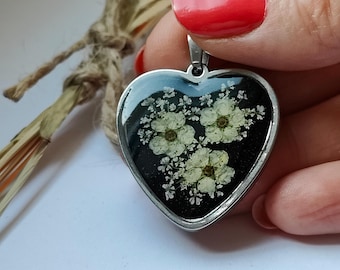 May include: A silver heart-shaped pendant with a black background and white pressed flowers inside.