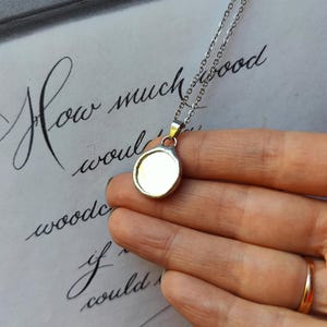 May include: A silver-colored pendant necklace with a round, blank face. The chain is thin and delicate. The background features handwritten script in black ink on a white surface.