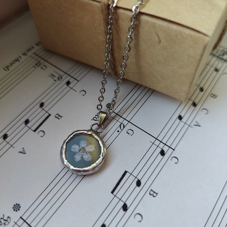 May include: A silver chain necklace with a round pendant featuring a white flower pressed inside clear resin.