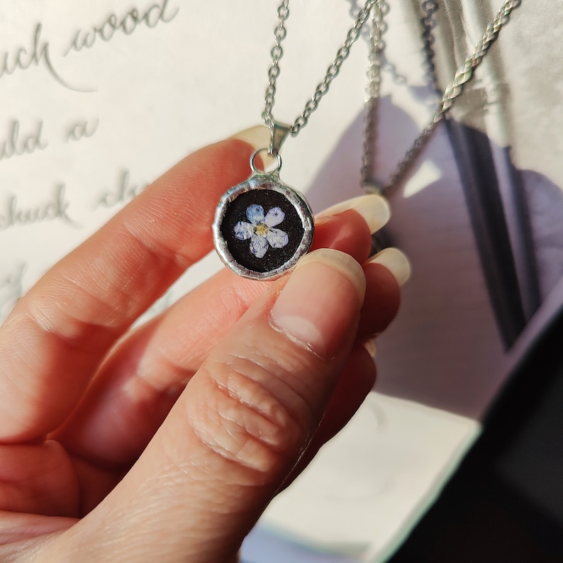 May include: A silver chain necklace with a round pendant featuring a pressed forget-me-not flower encased in resin.
