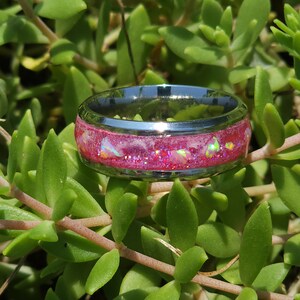 May include: A stainless steel ring with a pink and iridescent glitter inlay. The ring is set against a backdrop of green foliage.