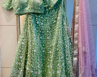 Op de afbeelding: Een groene geborduurde lehenga met een bijpassend groen geborduurd blouse. De lehenga heeft een bloemenpatroon met zilveren pailletten. De blouse heeft een V-hals en korte mouwen. De outfit wordt gecombineerd met een roze net dupatta met zilveren borduurwerk.