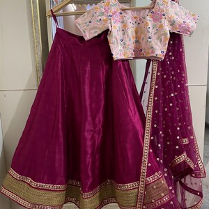 Puede incluir: Una falda de seda magenta con ribete dorado y magenta. La falda se combina con una blusa bordada rosa de manga corta. Un dupatta de red magenta con detalles dorados completa el atuendo.