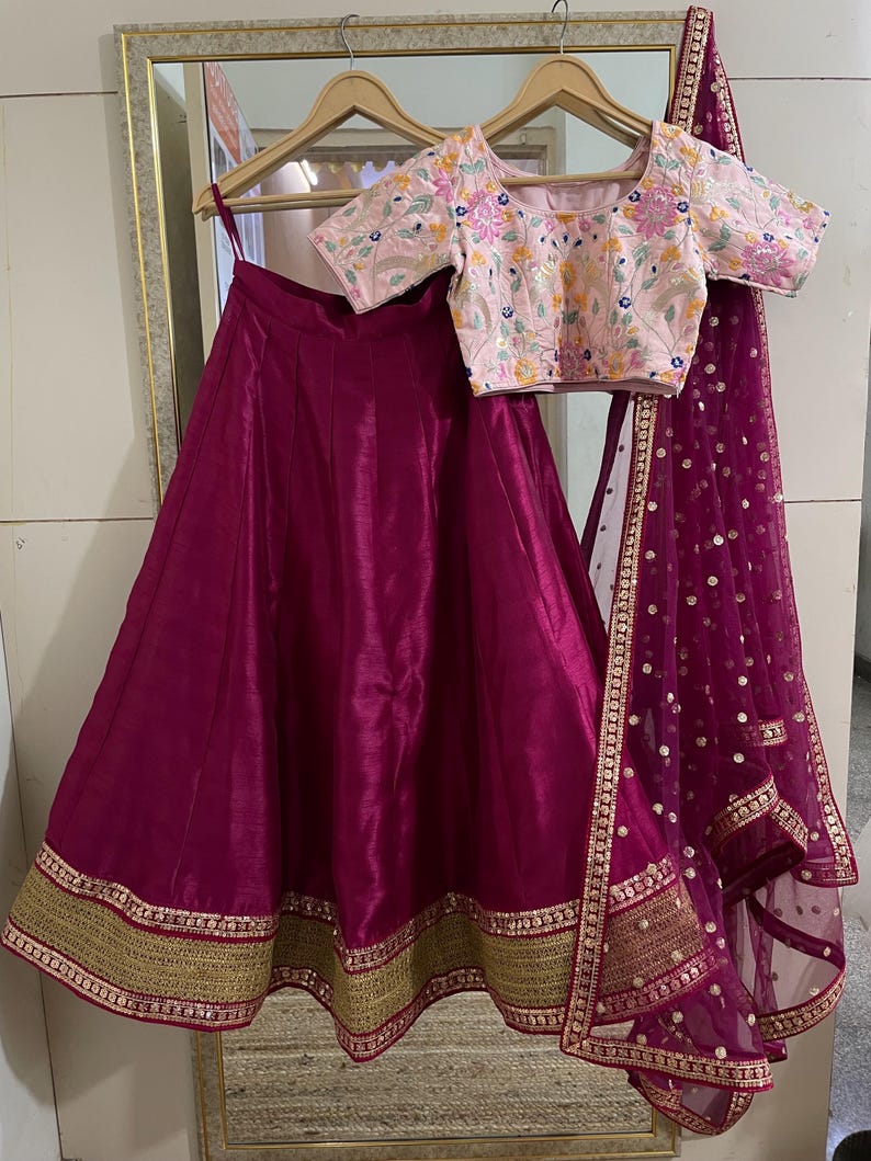 Puede incluir: Una falda de seda magenta con ribete dorado y magenta. La falda se combina con una blusa bordada rosa de manga corta. Un dupatta de red magenta con detalles dorados completa el atuendo.