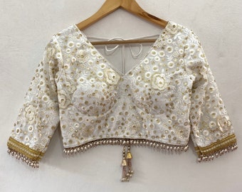Puede incluir: Blusa blanca bordada con hilo dorado y ribete de perlas. La blusa tiene un escote en V y mangas tres cuartos. Un borla de perlas cuelga de la parte inferior de la blusa.