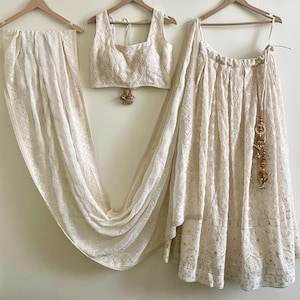 May include: A cream-colored, three-piece outfit, including a top, skirt, and scarf, hangs on wooden hangers. The fabric features an intricate, embroidered pattern. The skirt has a drawstring waist and a decorative tassel. The top is a cropped style.