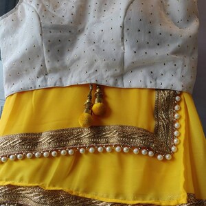 Pu&ograve; includere: Un sari giallo con una blusa bianca ricamata e bordi dorati. Il sari ha un bordo di perline con perle bianche.
