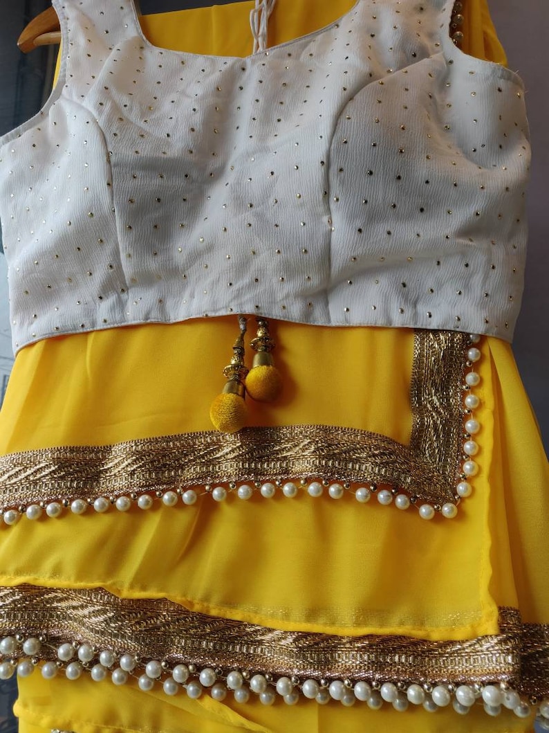 Pu&ograve; includere: Un sari giallo con una blusa bianca ricamata e bordi dorati. Il sari ha un bordo di perline con perle bianche.