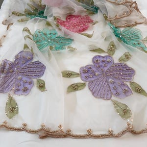 May include: White sheer fabric with embroidered floral designs in shades of purple, teal, and pink. The flowers are embellished with sequins and beads, and the edges are trimmed with gold-colored beads. Green leaves are also embroidered.