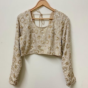 Könnte beinhalten: Ein beiges, langärmeliges Crop-Top mit aufwendigen goldenen und weißen Perlenarbeiten. Das Oberteil hat einen Rundhalsausschnitt und ist mit kreisförmigen Mustern aus Perlen und Pailletten verziert. Es hängt an einem Holzbügel vor einem neutralen Hintergrund.