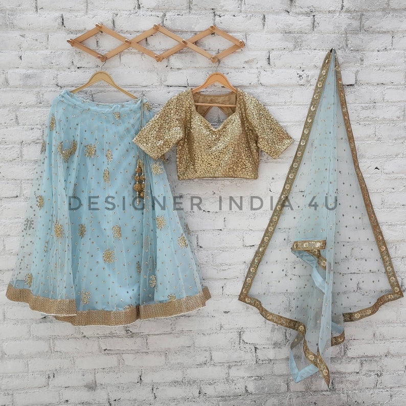 May include: A light blue embroidered lehenga with a gold embroidered blouse and a matching sheer dupatta. The outfit is displayed on hangers against a white brick wall.