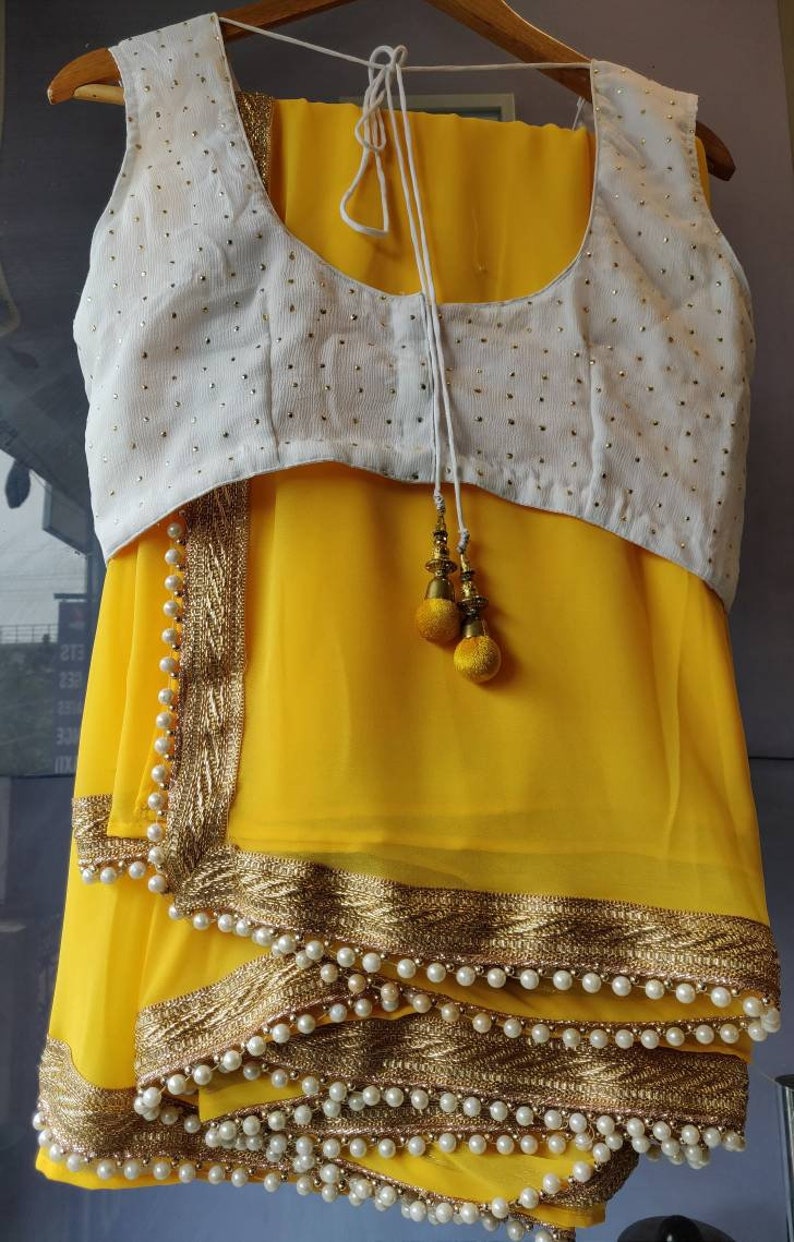 Pu&ograve; includere: Un sari di chiffon giallo con una blusa bianca ricamata. Il sari ha un bordo dorato e perlato.