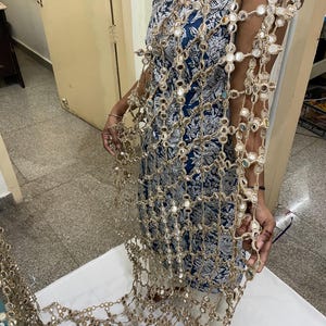 May include: A gold-colored, open-weave shawl with small, round mirrors attached throughout. The shawl is draped over a blue and white patterned dress. The mirrors reflect light, creating a shimmering effect.