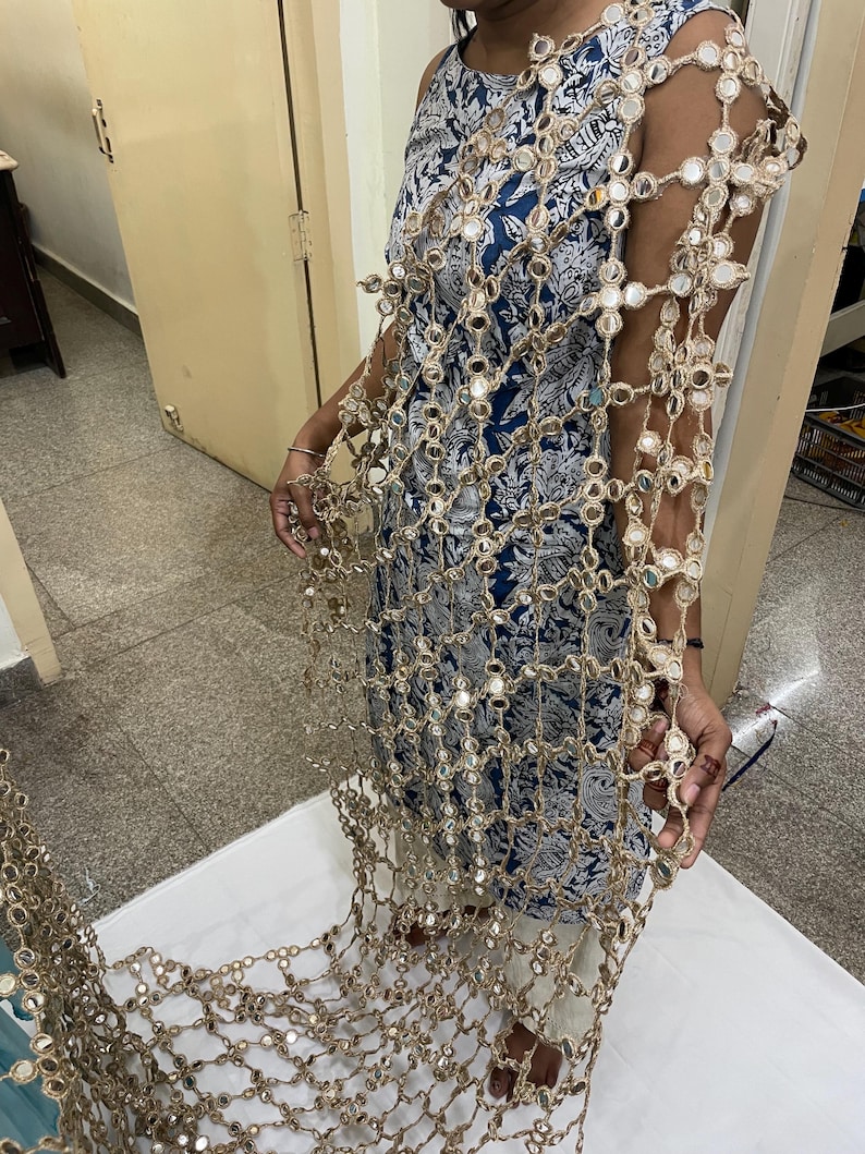 May include: A gold-colored, open-weave shawl with small, round mirrors attached throughout. The shawl is draped over a blue and white patterned dress. The mirrors reflect light, creating a shimmering effect.