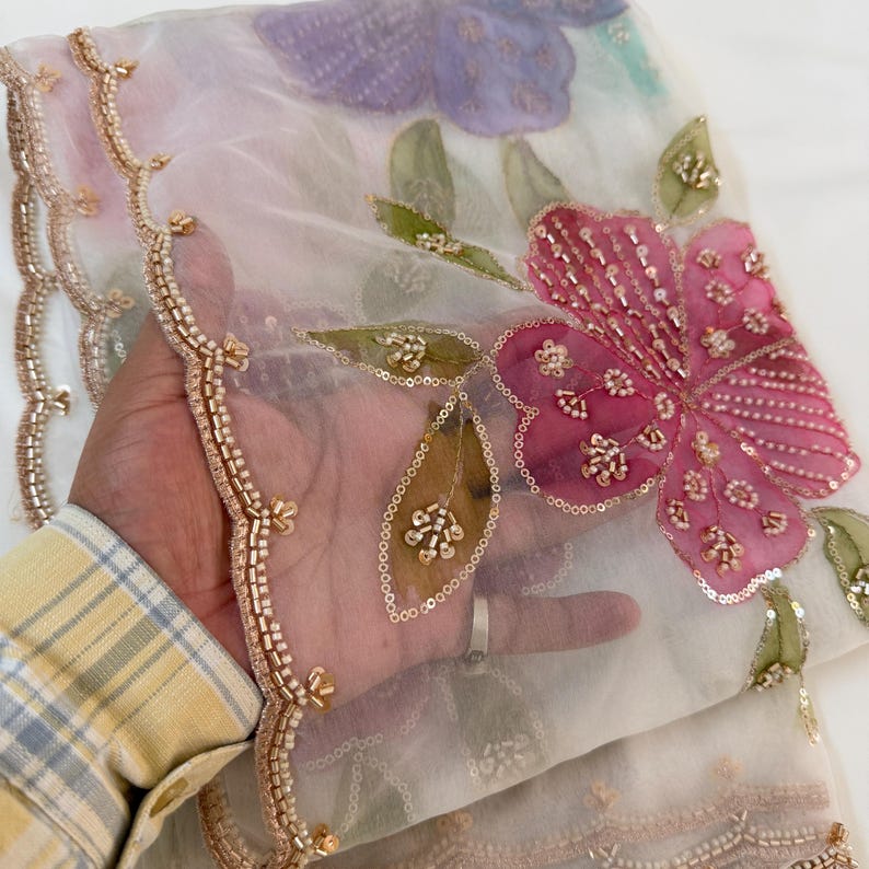 May include: Sheer, off-white fabric with embroidered floral designs. The fabric features a large pink flower with sequin and bead embellishments, along with green leaves. The edges are trimmed with gold-colored beads, creating a delicate and ornate appearance.