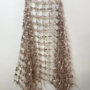 May include: A beige, open-weave textile with small, round mirrors attached at the intersections of the weave. The textile is hanging from a wooden hanger against a white wall. The mirrors reflect light, creating a shimmering effect.