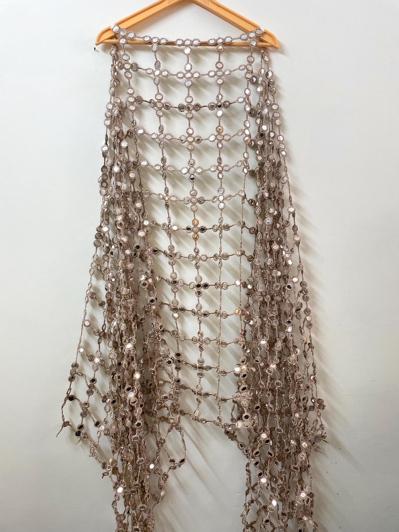 May include: A beige, open-weave textile with small, round mirrors attached at the intersections of the weave. The textile is hanging from a wooden hanger against a white wall. The mirrors reflect light, creating a shimmering effect.