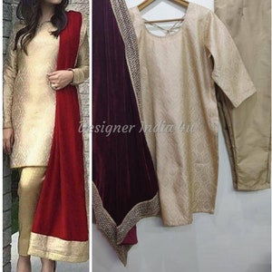 May include: A three-piece outfit in a gold color with a maroon velvet dupatta. The dupatta has a gold trim with embellishments. The outfit includes a long-sleeved top, pants, and a dupatta.