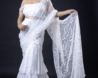 May include: A white saree with a sheer, embroidered dupatta. The saree has a layered skirt and a fitted bodice. The dupatta is decorated with sequins and floral patterns.
