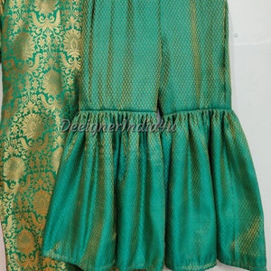 May include: A two-piece green and gold outfit. The top is a long-sleeved tunic with a floral pattern. The pants are wide-legged with a ruffled hem. The fabric has a textured appearance. The outfit is displayed on a hanger.