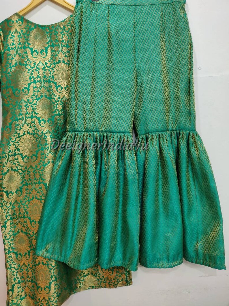 May include: A two-piece green and gold outfit. The top is a long-sleeved tunic with a floral pattern. The pants are wide-legged with a ruffled hem. The fabric has a textured appearance. The outfit is displayed on a hanger.
