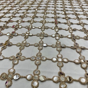 May include: Gold-colored, open-weave fabric with a grid pattern. Each intersection features a cluster of small, round, reflective mirrors. The fabric is likely used for decorative purposes, such as embellishing clothing or home decor.