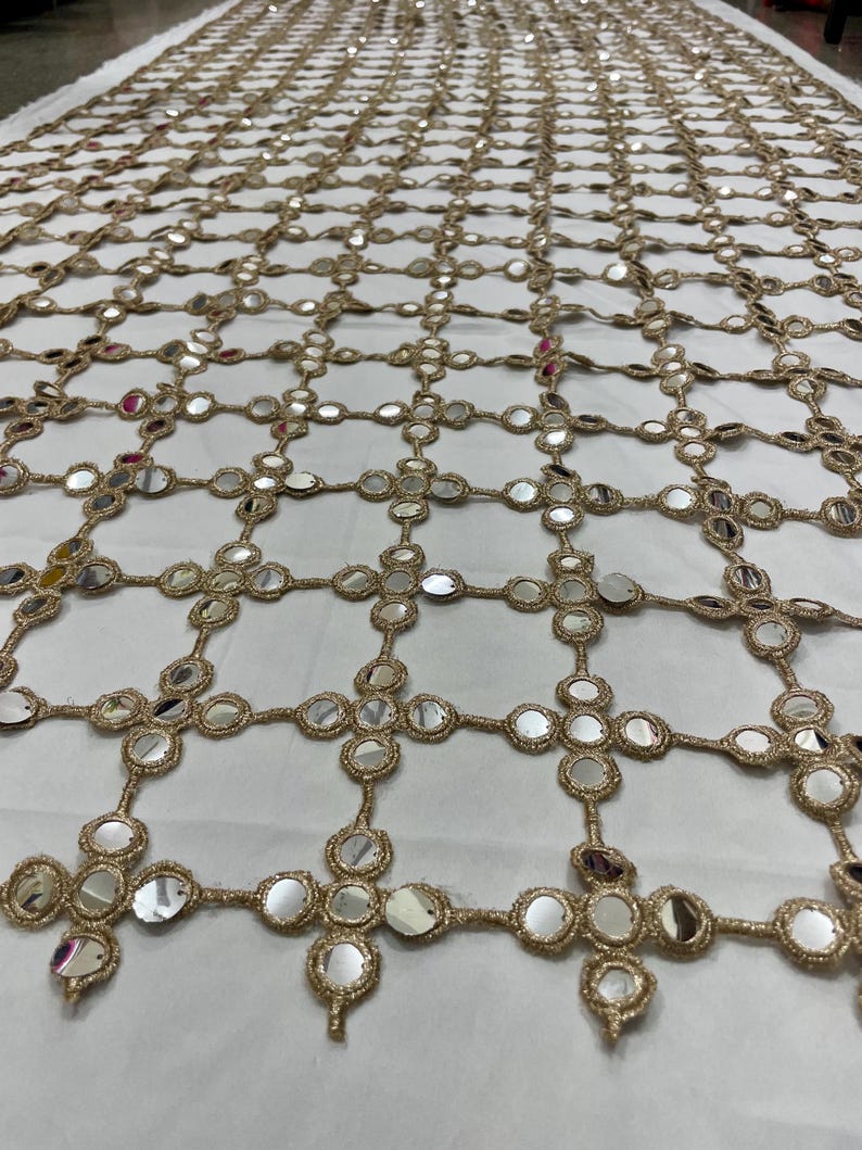 May include: Gold-colored, open-weave fabric with a grid pattern. Each intersection features a cluster of small, round, reflective mirrors. The fabric is likely used for decorative purposes, such as embellishing clothing or home decor.