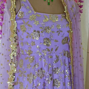 May include: A lavender-colored dress with gold floral embroidery and sequins. The dress has a sleeveless design and a sheer dupatta with gold trim.
