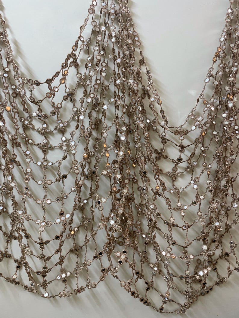 May include: A shimmering, draped accessory featuring a delicate net of interconnected strands. Each strand is adorned with small, reflective, circular embellishments, creating a cascade of light and texture. The color is a soft, warm brown.