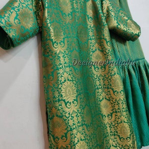 May include: A green and gold floral patterned outfit, including a tunic and flared pants. The fabric has a shiny, textured appearance. The tunic has short sleeves and a round neckline. The outfit is hanging on a wooden hanger.