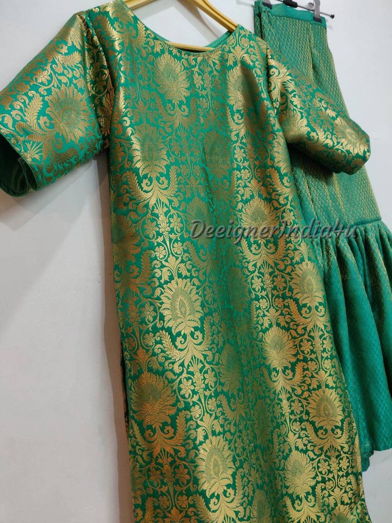 May include: A green and gold floral patterned outfit, including a tunic and flared pants. The fabric has a shiny, textured appearance. The tunic has short sleeves and a round neckline. The outfit is hanging on a wooden hanger.