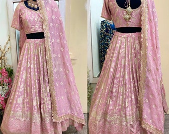 May include: A pink and gold embroidered lehenga with a matching embroidered blouse and a sheer dupatta. The lehenga has a flared skirt with a gold border and the blouse has a round neckline and short sleeves.