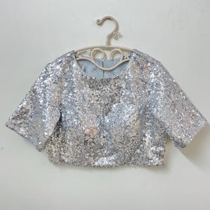 Puede incluir: Top corto plateado con lentejuelas y mangas cortas, colgado de una percha color crema. La parte superior está cubierta de brillantes lentejuelas plateadas. El escote y las mangas están cuidadosamente acabados, creando un look glamuroso.