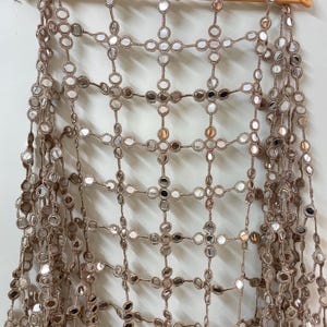 May include: A tan-colored, open-weave textile with small, round, mirrored accents. The textile is draped over a wooden hanger. The mirrored accents are evenly spaced throughout the design, creating a reflective pattern.