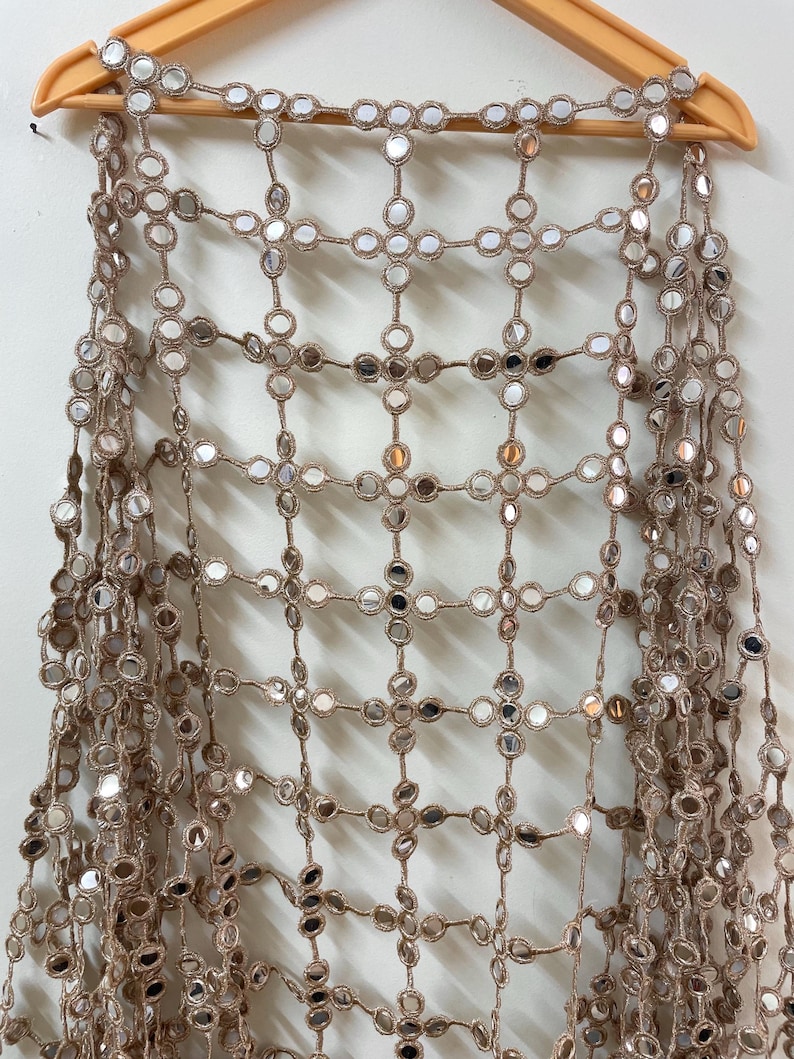 May include: A tan-colored, open-weave textile with small, round, mirrored accents. The textile is draped over a wooden hanger. The mirrored accents are evenly spaced throughout the design, creating a reflective pattern.