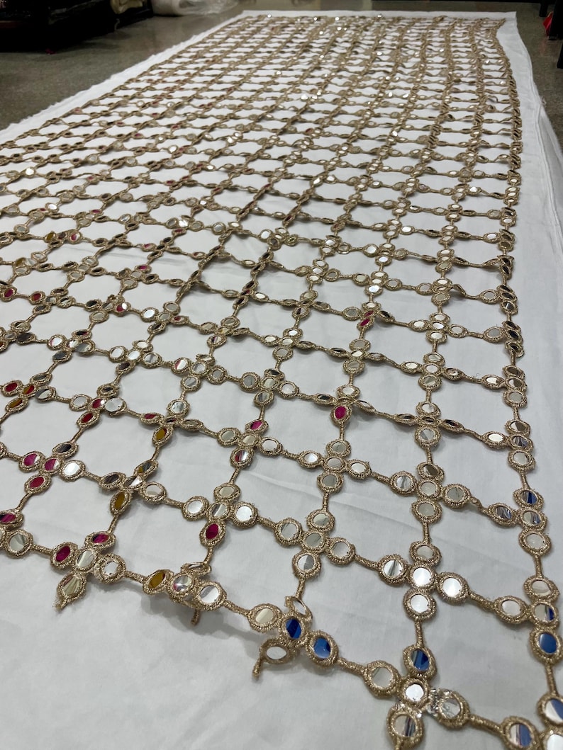 May include: A large, rectangular textile with a grid-like pattern. The grid is made of gold-colored thread, and each intersection features a small, round mirror. The mirrors reflect light and have colorful accents.