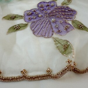 May include: Close-up of a white fabric with a large, embroidered purple flower and green leaves. The flower and leaves are embellished with beads and sequins. A gold beaded trim lines the bottom edge.