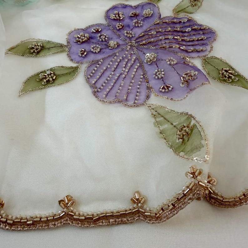 May include: Close-up of a white fabric with a large, embroidered purple flower and green leaves. The flower and leaves are embellished with beads and sequins. A gold beaded trim lines the bottom edge.