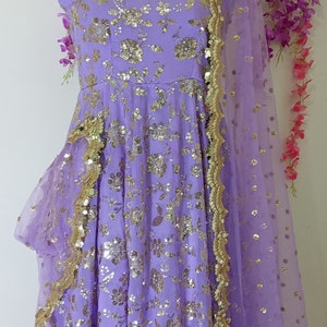 May include: A lavender-colored dress with gold sequins and floral embroidery. The dress has a ruffled hem and a matching sheer dupatta with a gold border.