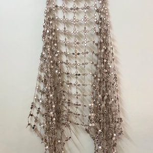 May include: A decorative, open-weave shawl with a grid pattern, adorned with numerous small, round, reflective mirror accents. The shawl is a neutral beige color and hangs from a wooden hanger against a white wall.