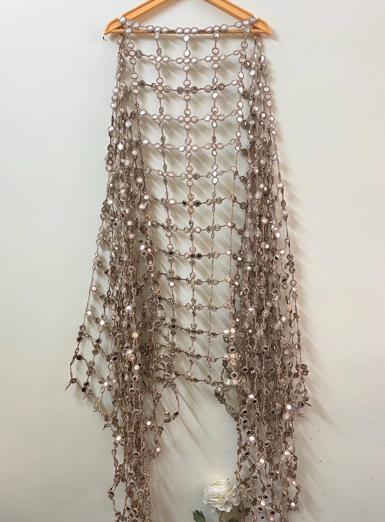 May include: A decorative, open-weave shawl with a grid pattern, adorned with numerous small, round, reflective mirror accents. The shawl is a neutral beige color and hangs from a wooden hanger against a white wall.