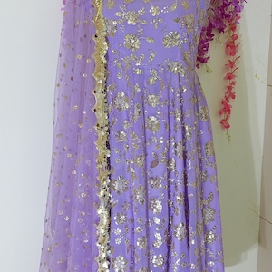 May include: A lavender-colored dress with gold sequins and a matching sheer dupatta. The dress has a square neckline and sleeveless design. The dupatta has a gold border and is decorated with sequins.