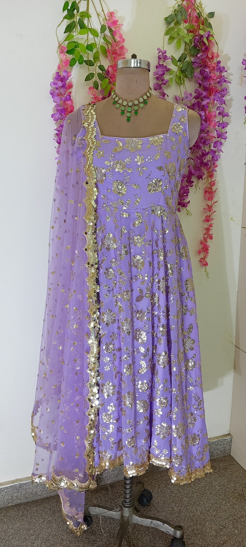 May include: A lavender-colored dress with gold sequins and a matching sheer dupatta. The dress has a square neckline and sleeveless design. The dupatta has a gold border and is decorated with sequins.